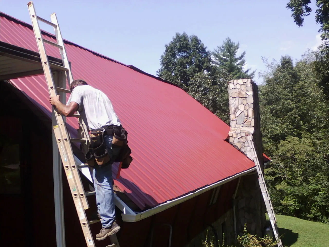 Roofer on a ladder beside a red metal roof and stone chimney — Soffit & Fascia Repair by Roofing Brothers in Paxtonville